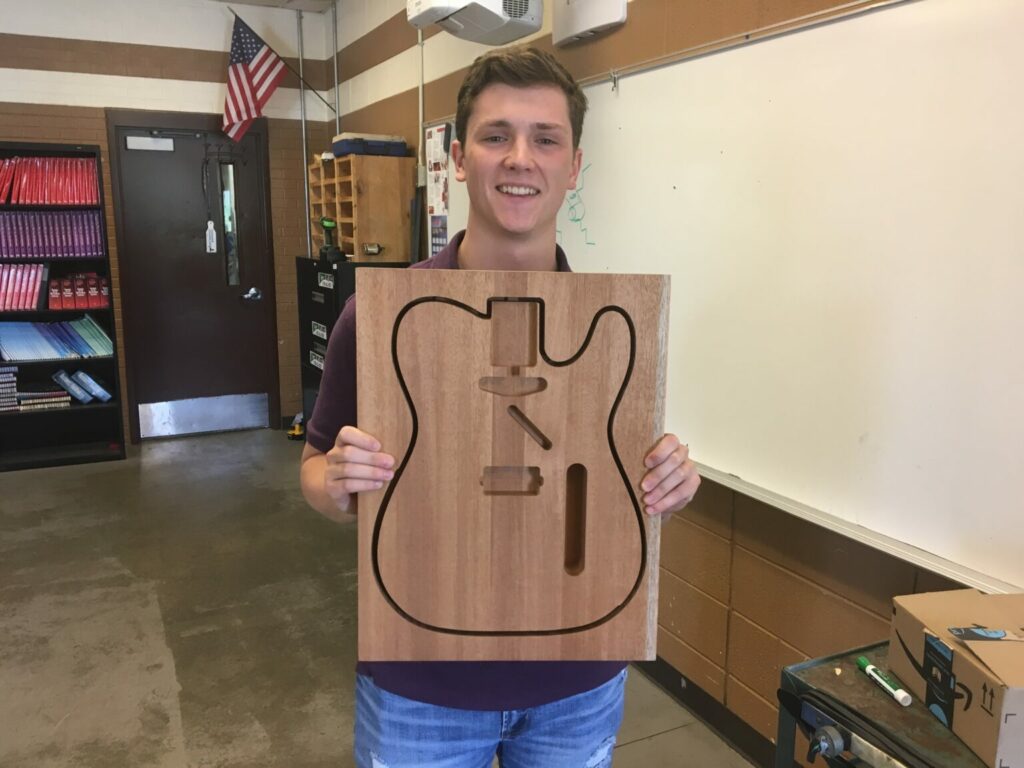 Blonde male student smiling holding wooden carving of electric guitar body inside a classroom