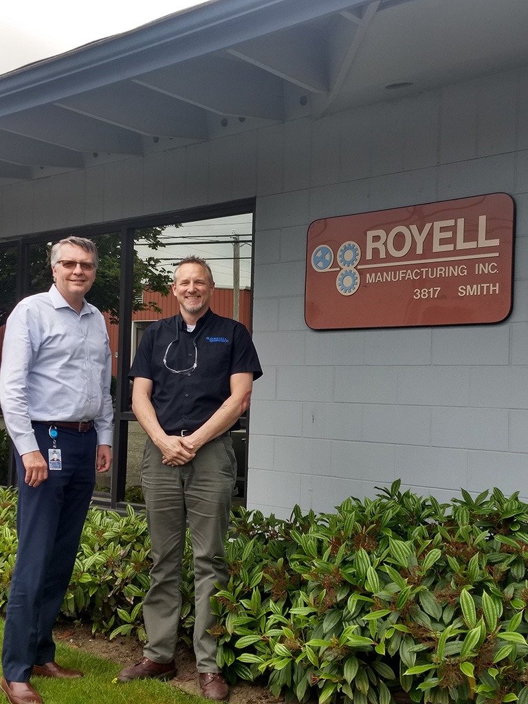 Two employees smiling in front of a building with a sign that reads Royal Manufacturing INC 3817 Smith